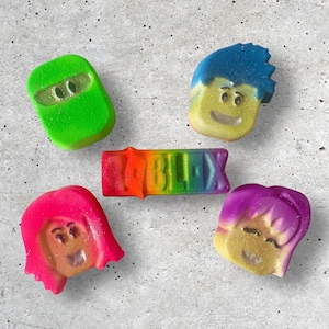Może przedstawiać: Pięć kolorowych bombek do kąpieli w kształcie postaci z Roblox. Bombki do kąpieli są zielone, niebieskie, różowe, fioletowe i tęczowe. Tęczowa bombka do kąpieli ma napisane na niej "Roblox".