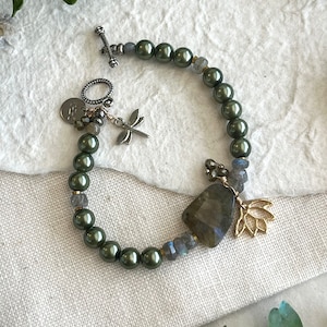 May include: A green pearl bracelet with a dragonfly charm, a large labradorite stone, and a gold lotus charm. The bracelet has a silver toggle clasp.