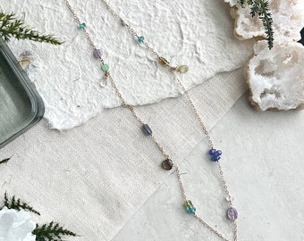 May include: A delicate gold necklace with multiple strands, adorned with colorful gemstones in shades of blue, green, purple, and yellow. The necklace features a small gold charm and a teardrop-shaped pendant.