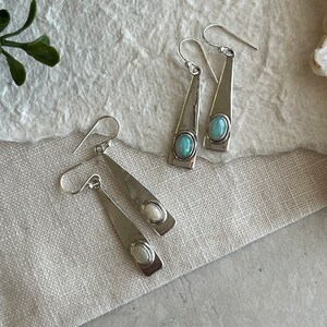 Puede incluir: Pendientes colgantes plateados con diseño triangular y gemas ovaladas iridiscentes. Los pendientes se exhiben sobre una superficie texturizada blanquecina, mostrando su acabado pulido y estilo elegante. Joyería adecuada para cualquier ocasión.