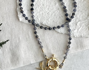 Op de afbeelding: Een ketting met donkergrijze kralen, een gouden sluiting en bedels. De ketting heeft een goudkleurige libellenbedel, een blauwgrijs kristal en een witte parel hanger. De sluiting is een grote, goudkleurige ring.