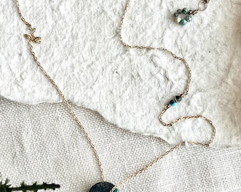 May include: A gold-colored necklace with a dark, round pendant featuring turquoise accents. The necklace has a delicate chain and includes turquoise beads and a safety pin clasp. A small gold disc with text is also present.