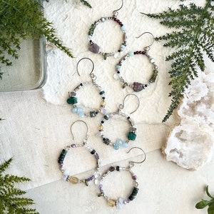 May include: Six pairs of hoop earrings with various semi-precious stones and beads. The earrings are silver-toned and feature a variety of colors, including green, blue, purple, and brown.