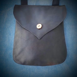 May include: A dark blue leather pouch with a flap closure and a button. The pouch has a rounded bottom and two straps. The button is a light tan color. The background is a textured blue.