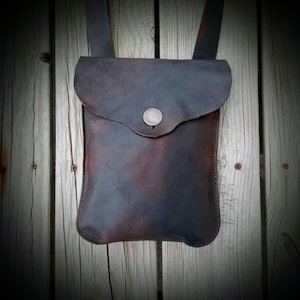 May include: A dark brown leather pouch with a flap closure secured by a single silver-toned metal snap button. The bag features a rectangular body with rounded bottom corners and a long, wide leather strap extending from the top for carrying.