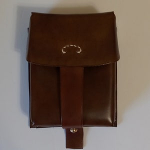 May include: A brown leather pouch with a flap and a white stitched design. The pouch has a strap with a brass button.