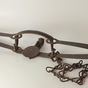 May include: A vintage metal animal trap with a circular plate, chain, and hinged arms. The trap is made of dark, weathered metal, suggesting age and use. The chain is attached to the trap's base.