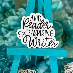 May include: A white sticker with black cursive text that reads "AVID Reader ASPIRING Writer." The sticker is displayed on a small, light blue wooden easel. The background features a blurred arrangement of blue flowers and a marbled surface.