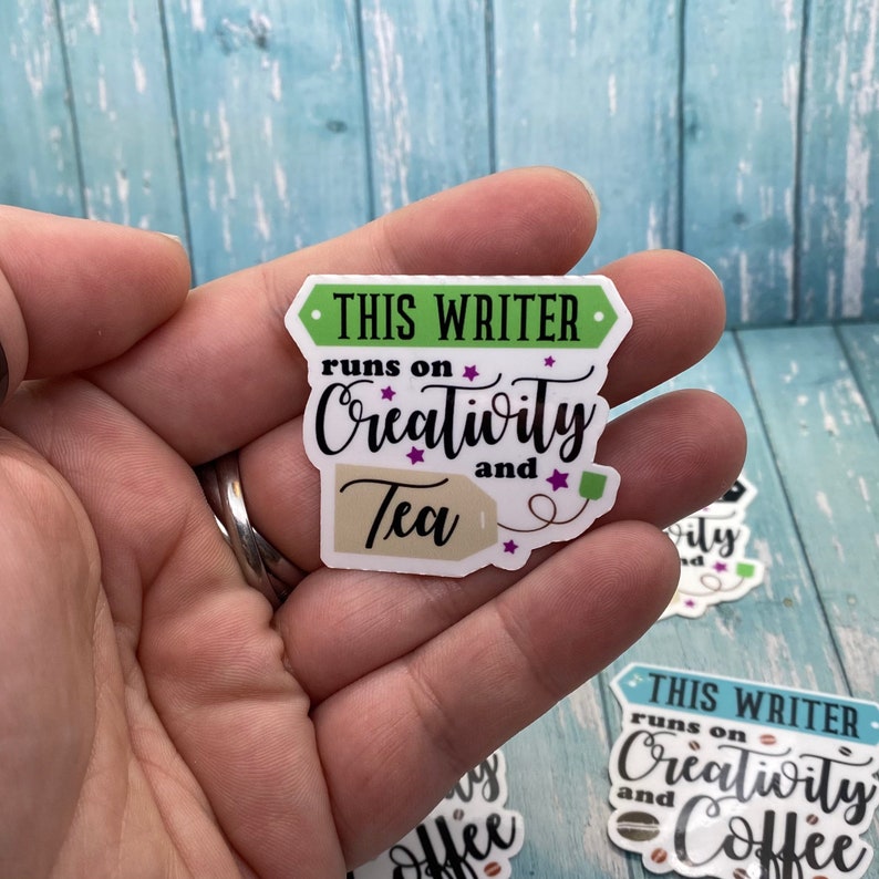 May include: A sticker with the text "THIS WRITER runs on Creativity and Tea" with a tea bag tag. The sticker is white with green and purple accents.