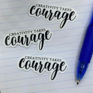 May include: Three white stickers with black text that reads "Creativity Takes Courage" on a lined notebook page. A blue pen is lying on the page.