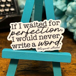 May include: White sticker with black text that reads "If I waited for perfection, I would never write a word."  The text is in a cursive font.  The sticker is on a teal easel.