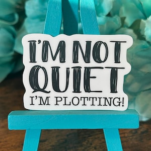 May include: White sticker with black text that reads "I'm not quiet I'm plotting!" The sticker is displayed on a small turquoise easel.