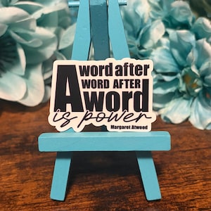 May include: A white sticker with black text that reads "A word after word after word is power" by Margaret Atwood. The sticker is displayed on a small, light blue wooden easel, with a blurred floral background.