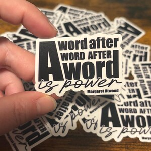 May include: A white sticker with black text that reads "A word after word after word is power" with "Margaret Atwood" below. The sticker is held by a hand, with other similar stickers in the background.