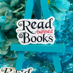 Könnte beinhalten: Weißer Aufkleber mit schwarzem Text, der "Read banned Books" lautet, auf einem kleinen blauen Staffelei. Der Aufkleber hat einen abgenutzten Look mit roten Spritzern.
