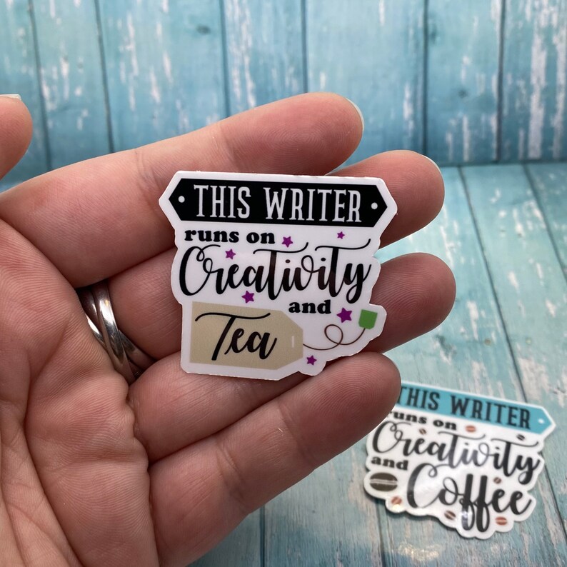 May include: A white sticker with black text that reads "This Writer runs on Creativity and Tea". The sticker features a brown tea bag with a white string and a green tag. The sticker has purple stars around the text.