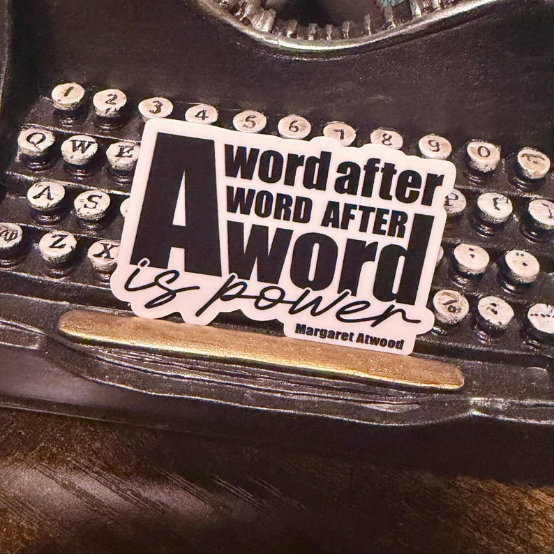 May include: A sticker with the text "A word after word after word is power" in a bold, black font. The sticker is placed on a vintage typewriter, with the keys and body of the machine visible.