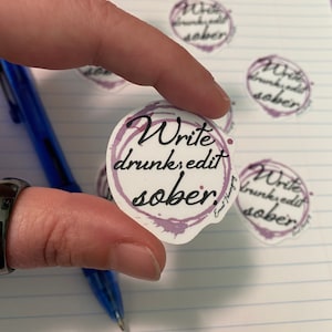 Puede incluir: Una pegatina blanca con el texto "Write drunk, edit sober" en escritura negra, rodeada por un diseño de acuarela púrpura. Varias pegatinas similares son visibles en una superficie de papel rayado.