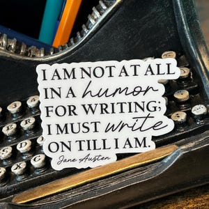 May include: A white sticker with black text that reads "I am not at all in a humour for writing; I must write on till I am."  The text is attributed to Jane Austen. The sticker is placed on a vintage typewriter.