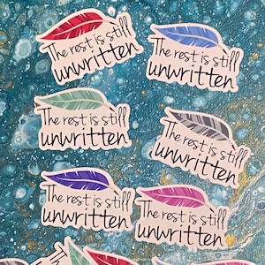 Puede incluir: Seis pegatinas de plumas de colores con el texto "The rest is still unwritten" en cada una. Las pegatinas están sobre un fondo azul.