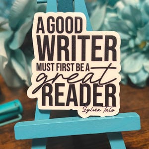 May include: A white sticker with black text that reads "A GOOD WRITER MUST FIRST BE A great READER" is displayed on a small blue easel. The sticker has a scalloped edge and the name "Sylva Talo" is written at the bottom. The background is blurred.