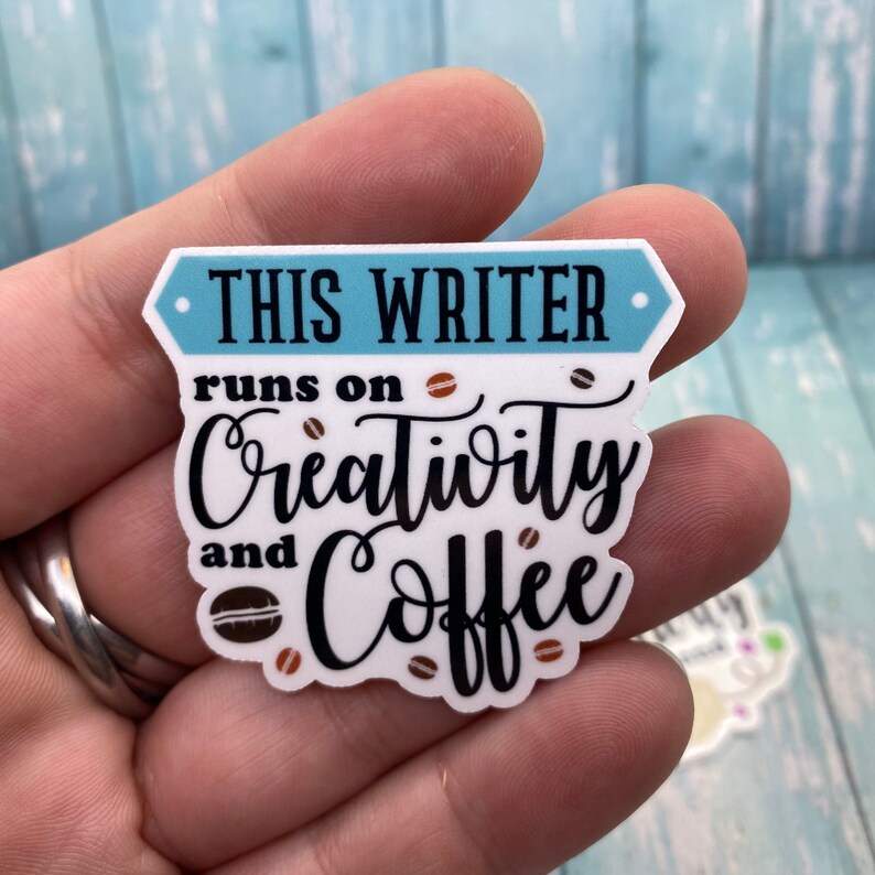 May include: White sticker with black text that reads "This writer runs on creativity and coffee". The sticker has a blue border and brown coffee bean illustrations.