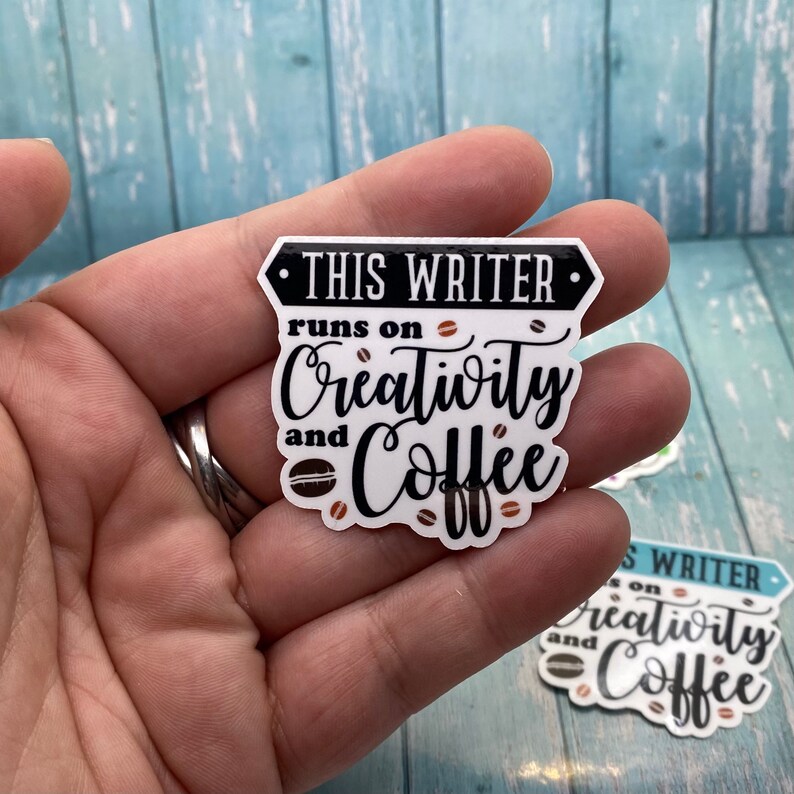 May include: White sticker with black text that reads "This Writer runs on Creativity and Coffee". The sticker has a black border and a coffee bean design.