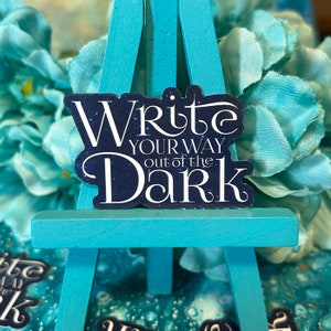 May include: A blue and white sticker with the text "Write your way out of the dark" on a teal easel.