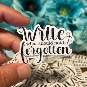 Puede incluir: Una pegatina blanca con texto negro que dice "Escribe lo que no debe ser olvidado" de Isabel Allende. La pegatina está sujeta por una mano, con un fondo borroso de flores azules y otras pegatinas.