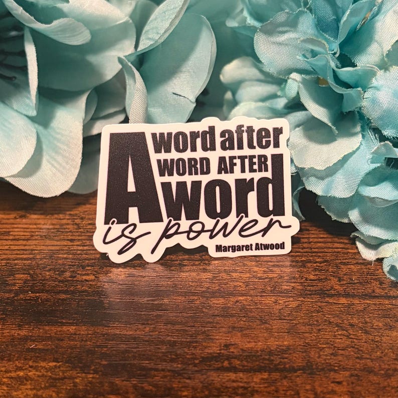 May include: A rectangular sticker with the text "A word after word after word is power" in black and white. The author's name, Margaret Atwood, is at the bottom. The sticker is on a wooden surface with blue flowers in the background.
