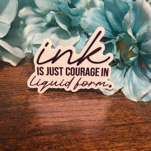May include: A white sticker with black text that reads "ink IS JUST COURAGE IN liquid form." The sticker has a black border and is placed on a wooden surface with blue flowers in the background.