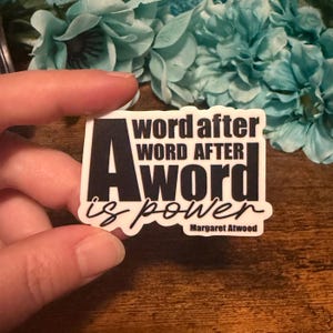 May include: A rectangular sticker with the text "A word after word after word is power" in black and white. The sticker also includes the author's name, Margaret Atwood, in a smaller font.