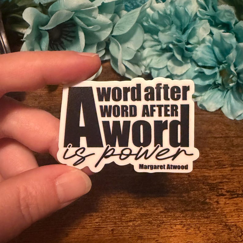 May include: A rectangular sticker with the text "A word after word after word is power" in black and white. The sticker also includes the author's name, Margaret Atwood, in a smaller font.