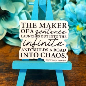 Könnte beinhalten: Ein weißer Aufkleber mit schwarzem Text auf einer kleinen blauen Staffelei. Der Text lautet: "THE MAKER OF A sentence LAUNCHES OUT INTO THE infinite AND BUILDS A ROAD INTO CHAOS." Das Zitat wird Ralph Waldo Emerson zugeschrieben.