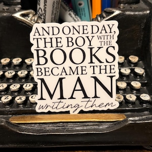 May include: A white sticker with black text that reads "AND ONE DAY, THE BOY WITH THE BOOKS BECAME THE MAN writing them." The sticker is placed on a vintage typewriter.