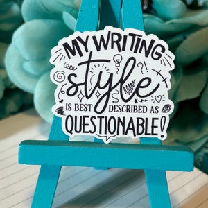 May include: A white sticker with black text that reads "MY WRITING STYLE IS BEST DESCRIBED AS QUESTIONABLE!" The sticker is on a small, turquoise wooden easel. The background is a blurred teal color.