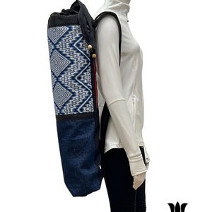 Peut inclure: Un sac de tapis de yoga avec un motif géométrique bleu et blanc sur la partie supérieure et un fond en jean. Le sac a des sangles noires et des accents de perles en bois. Le sac est porté sur le dos d'un mannequin.