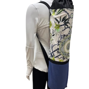 May include: A yoga mat bag with a black drawstring top and a navy blue bottom. The main body of the bag features a cream-colored fabric with a floral and geometric pattern in shades of green, blue, and gray. The bag has a black shoulder strap.