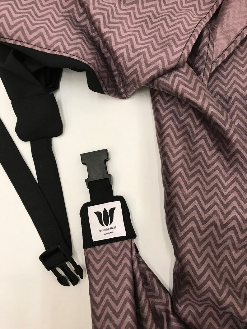 May include: A black and gray baby carrier with a zig-zag pattern. The carrier has a black strap with a buckle and a white label that says "My Yoga Room".