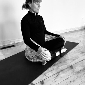 May include: A woman sits in a meditation pose on a black yoga mat, wearing a black long-sleeved shirt and a patterned fabric wrapped around her legs.