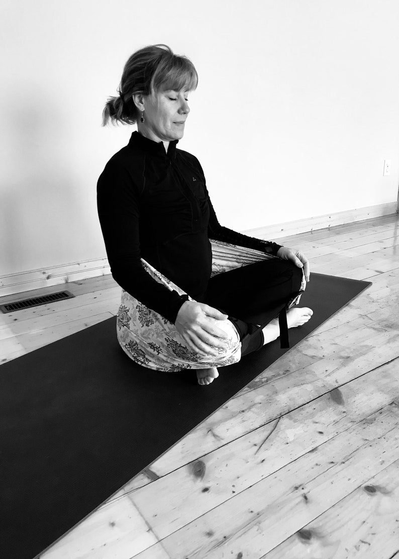 May include: A woman sits in a meditation pose on a black yoga mat, wearing a black long-sleeved shirt and a patterned fabric wrapped around her legs.