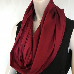 May include: A burgundy infinity scarf made of a soft, stretchy fabric. The scarf is worn around the neck and drapes down the front of the chest.