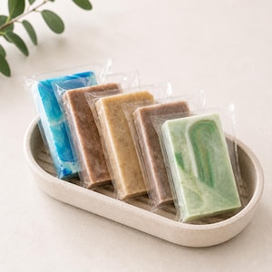 May include: Assortment of wrapped soap bars in various colors, including blue, brown, tan, and green, arranged in a beige ceramic dish. The soaps have unique marbled and layered designs. A sprig of green leaves is in the background.