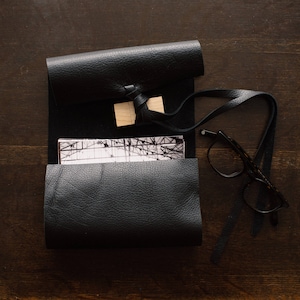 May include: A black leather roll-up case with a strap and a wooden accent. Inside, a black and white print is visible. A pair of black-framed glasses rests beside the case on a dark wooden surface. The case is open, revealing its contents.