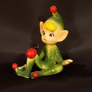May include: A ceramic figurine of a seated elf with a green outfit and hat, yellow hair, and a surprised expression. A red and black ladybug sits on the elf's leg. The figurine has red accents on the hat and feet.