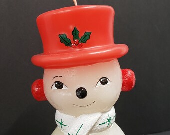 May include: A white snowman candle with a red hat, green holly, and a white scarf. The snowman is holding a candy cane in its right hand and has black eyes, a black nose, and three black buttons.