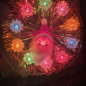May include: A circular holiday decoration featuring a central Santa Claus face with a white beard and pink hat, surrounded by gold metallic tinsel and eight star-shaped light covers in various colors including pink, blue, green, and orange.