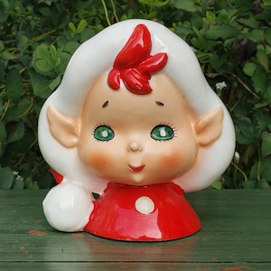 May include: A ceramic figurine of a smiling elf with green eyes, rosy cheeks, and a red bow. The elf wears a red dress with a white button and has white hair. A white ball is on the left side of the elf.