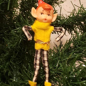 May include: A vintage Christmas elf ornament. The elf has a painted face with large ears, a red hat, and a yellow sweater. The arms and legs are made of fabric with a black and white checkered pattern. The elf is wearing yellow shoes.