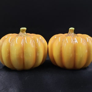 May include: Two ceramic pumpkin-shaped salt and pepper shakers. The shakers are a light orange color with a glossy finish.